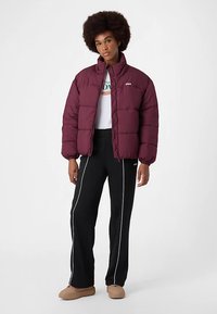 Bourgognefarvet dunjakke med høj krave og syede paneler, båret over en hvid grafik T-shirt og sorte træningsbukser med hvide striber.