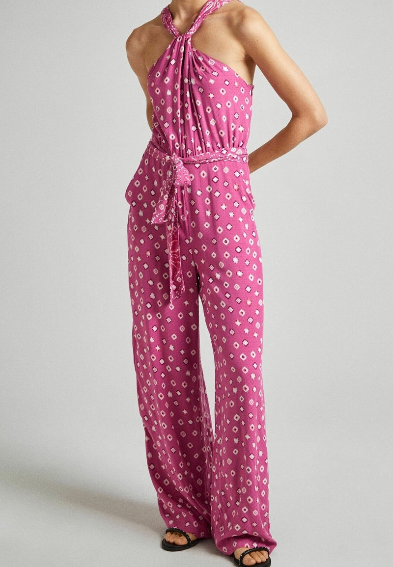 Combinaison rose avec un col halter, des jambes larges et une taille nouée, ornée de motifs blancs en forme de diamants et d'étoiles sur un tissu texturé.
