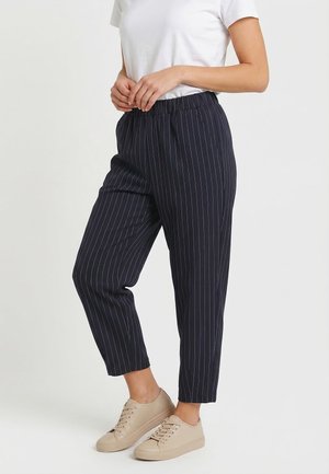 Persona vistiendo pantalones tobilleros negros de rayas, camiseta blanca y zapatillas beige, de pie con las manos entrelazadas al frente.