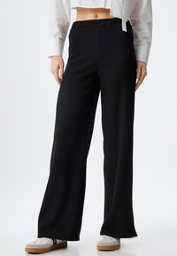 Zwarte wijde broek van textuurstof, met een elastische tailleband en een vloerlange snit. Draagt witte schoenen.