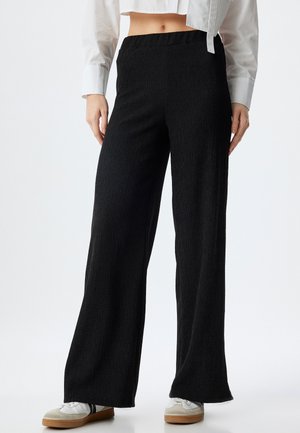 Zwarte wijde broek van textuurstof, met een elastische tailleband en een vloerlange snit. Draagt witte schoenen.