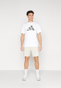 T-shirt blanche à manches courtes avec logo adidas camouflage noir et vert, associé à un short beige clair arborant un marquage adidas blanc.