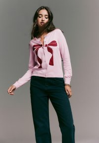 Light pink cardigan featuring a large burgundy bow design, made of soft knit fabric, with a button-up front and long sleeves.
