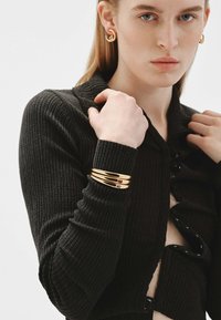 Cárdigan de punto negro acanalado con un cuello profundo, combinado con una pulsera de múltiples bandas doradas y pendientes redondos dorados, que destacan los acentos brillantes.