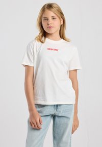 Camiseta de algodón blanca con el texto rojo "FRESH DAILY" en el pecho, con un corte holgado y mangas cortas. Combinada con unos vaqueros de color azul claro.