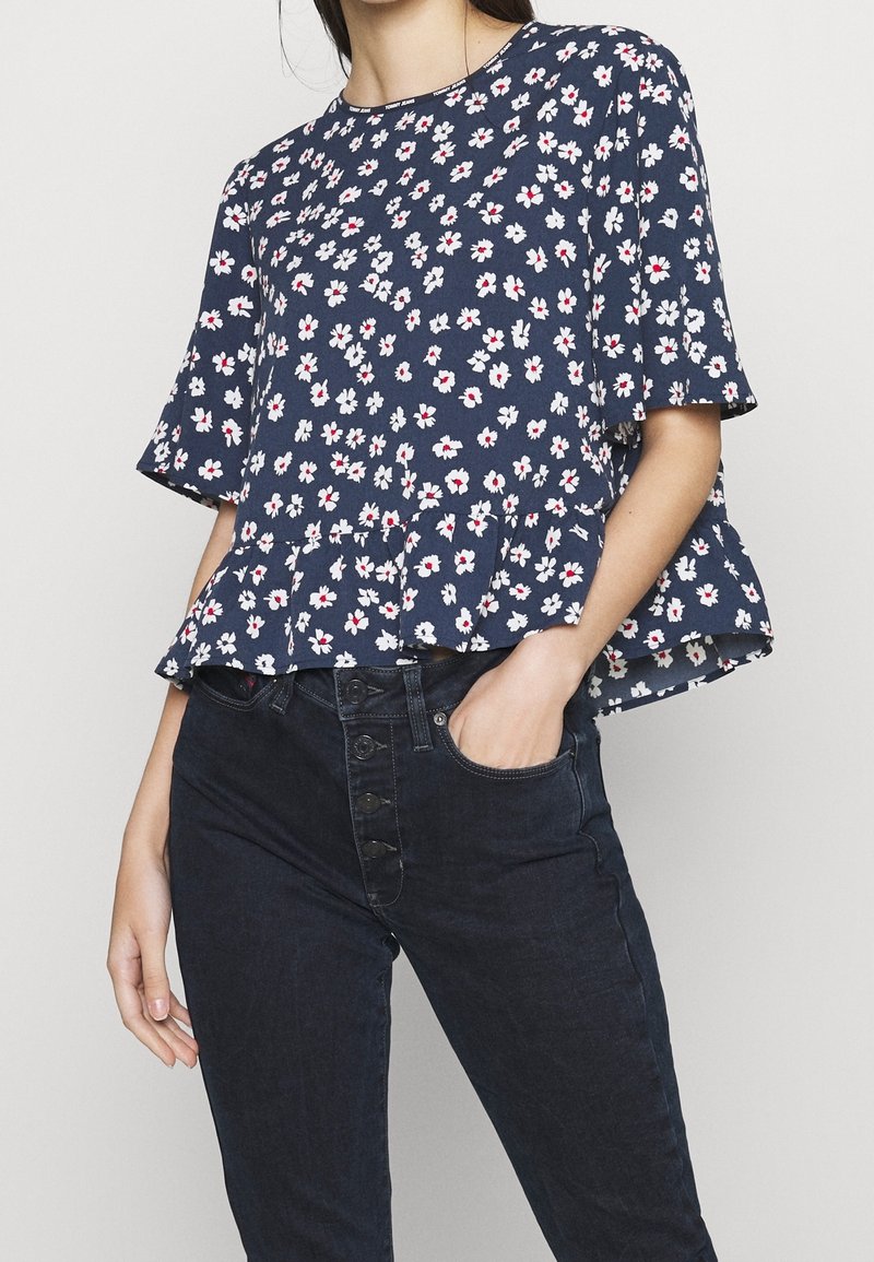 Navy blue floral peplum top featuring short sleeves, white and red flower patterns, and a round neckline, paired with dark denim pants.