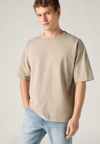 Μπεζ κοντομάνικη μπλούζα από βαμβάκι, με στρογγυλή λαιμόκοψη και μικρό λογότυπο στο στήθος, φορεμένη με ανοιχτόχρωμο τζιν παντελόνι.