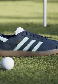 Ναυτικό μπλε αθλητικό παπούτσι Adidas Gazelle με λευκές ρίγες σε γρασίδι κοντά σε μια λευκή μπάλα του γκολφ και μια θολή σημαία τρύπας γκολφ στο φόντο.