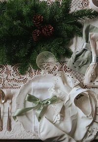 Hvid silikonebibs med blonder, grøn bånd, beige og grønne servietter, glas, og kogler på en blondeborddug med grøntstænger.