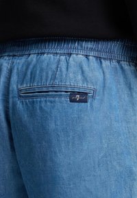 Pantaloni in denim azzurro chiaro a vestibilità rilassata con cintura elasticizzata, dotati di una tasca posteriore e di un'etichetta del marchio intrecciata cucita sopra la tasca.