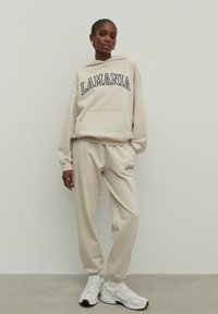 Beige kapucnis pulóver "LAMANIA" felirattal, elülső zsebbel, összeillő melegítőnadrággal. Sima anyag és laza szabás, fehér sportcipőkkel kiegészítve.