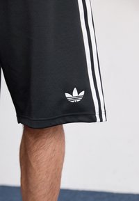 Sorte atletiske shorts lavet af blødt stof med hvide sidelister og et hvidt Adidas-logo nederst til venstre på benet.