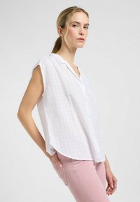 Blusa blanca de botones de manga corta con un patrón de cuadros texturizado, escote en V y dobladillo curvado, combinada con pantalones rosa claro.
