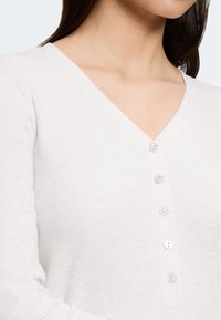 Donna che indossa un top bianco a costine con scollo a V e cinque bottoni trasparenti sul davanti, che mostra parte del collo e della spalla.