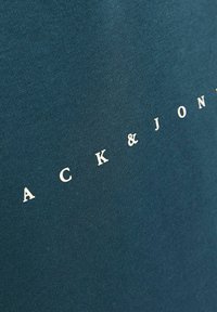 Camiseta de algodón azul con texto blanco que dice "ACK & JONE". Textura suave, diseño de cuello redondo, vista en primer plano que resalta los detalles de la tela.