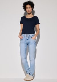 Camiseta de manga corta en azul marino oscuro combinada con jeans ajustados de color azul claro y zapatillas crema. El modelo está de pie con las manos en los bolsillos contra un fondo neutro.