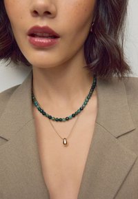 Zwei geschichtete Halsketten: eine mit grünen und blauen Edelsteinperlen, die andere mit einem goldenen Tropfenanhänger. Getragen über einem beigen Blazer.