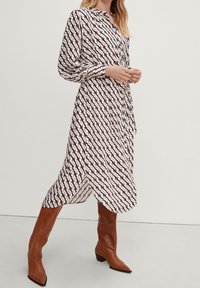 Frau trägt ein gemustertes, langärmliges Midi-Kleid mit Knöpfen und braunen knielangen Spitzenschuhen und steht vor einem schlichten Hintergrund.