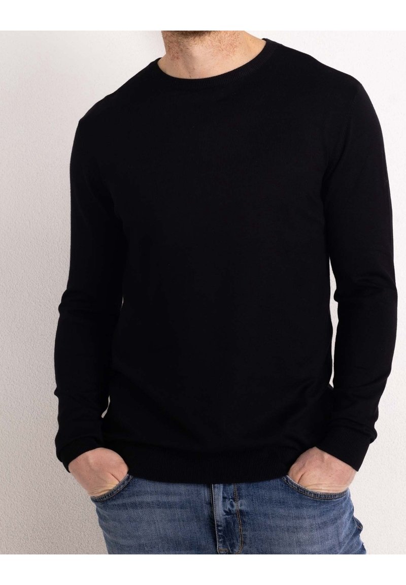 Uomo con un maglione nero a maniche lunghe e collo rotondo, con le mani nelle tasche dei jeans blu, su uno sfondo bianco semplice.