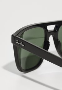 Ochelari de soare din plastic negru cu lentile verzi, având o formă rectangulară îndrăzneață și logo-ul Ray-Ban pe braț.