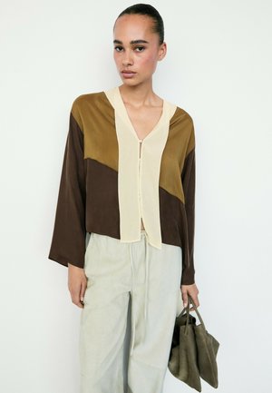 Persona con i capelli raccolti in uno chignon indossa una camicetta color block marrone, beige e crema, pantaloni chiari e tiene una morbida borsa verde oliva. Sfondo neutro.