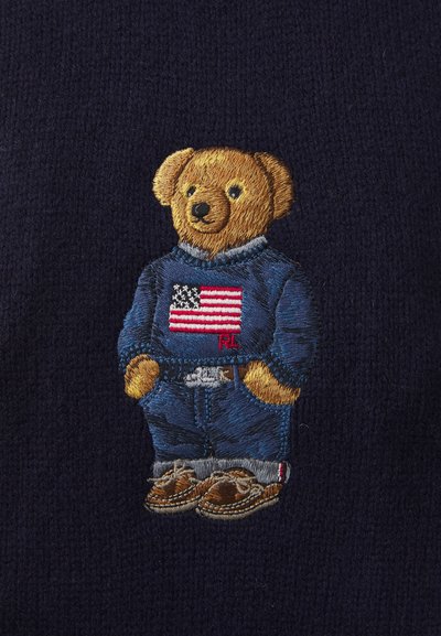 Suéter de color navy con un oso bordado que lleva una camisa azul con una bandera americana, unos vaqueros de mezclilla y zapatos marrones, con detalles texturizados.