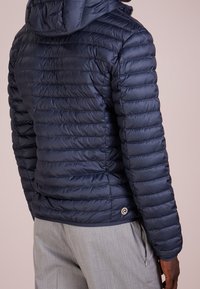 Marinblå puffjacka med horisontellt quiltat mönster, huva och elastiska mudd. Har en cirkulär logotyp vid fållen.