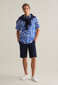 Camisa azul de manga corta con patrón floral blanco, combinada con pantalones cortos azul marino. Un suéter azul marino está drapeado sobre los hombros, con zapatillas blancas.