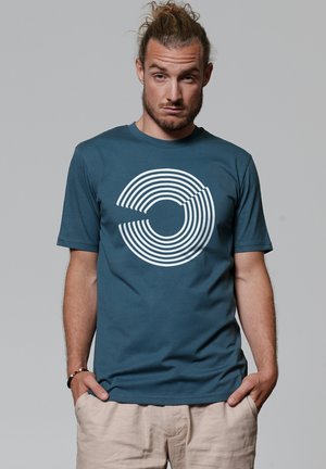 Teal cotton t-shirt with a large white circular line pattern on the front. Short sleeves and crew neck design, paired with beige shorts.
