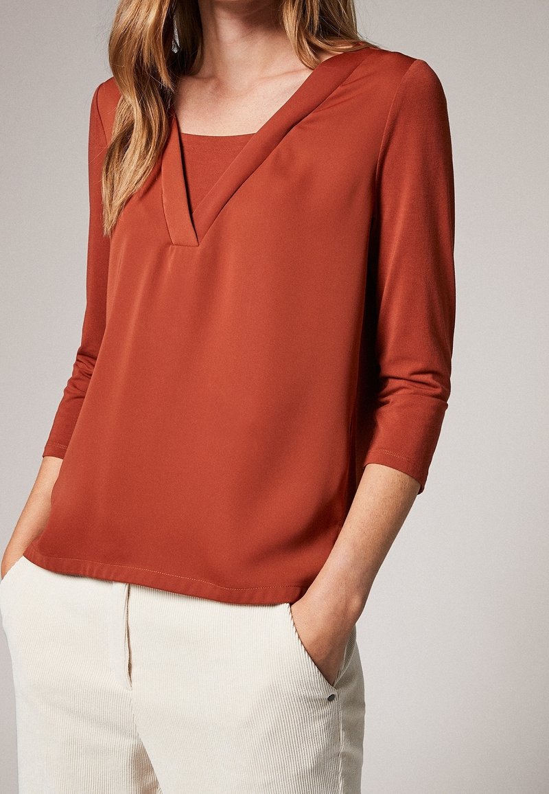 Top color ruggine con scollo a V e maniche a tre quarti, realizzato in tessuto morbido. Abbinato a pantaloni in corduroy color crema.