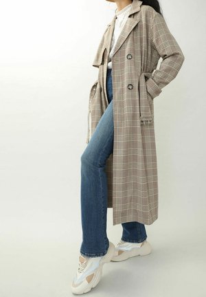 Femme portant un long trench-coat beige à carreaux, un haut en dentelle blanche, un jean bleu et des baskets blanches épaisses, debout devant un fond uni.