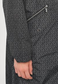 Jas van zwarte stof met een herhalend wit logo-patroon, voorzien van een ritssluitingzak en een gladde textuur. Geaccentueerd met een zilveren knoop.