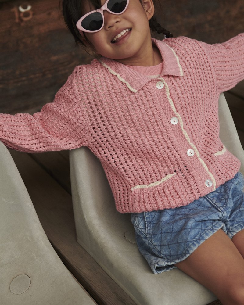 Junges Mädchen trägt eine rosa gestrickte Strickjacke und rosa Sonnenbrille, sitzt und lächelt mit ausgestreckten Armen auf beigen Sitzen.