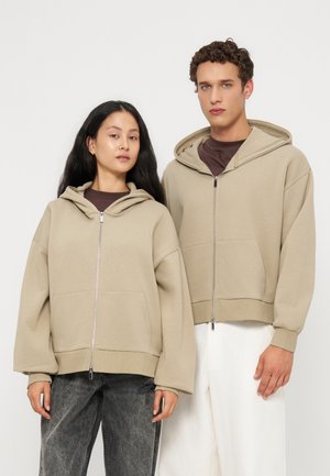 Deux jeunes adultes portant des sweats à capuche beige zippés, l’un avec un jean gris foncé et l’autre avec un pantalon blanc, debout devant un fond uni.