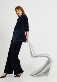 Costume en velours bleu marine avec blazer croisé et pantalon à jambes larges, accompagné d'une chaise argentée élégante et de talons pointus marron.