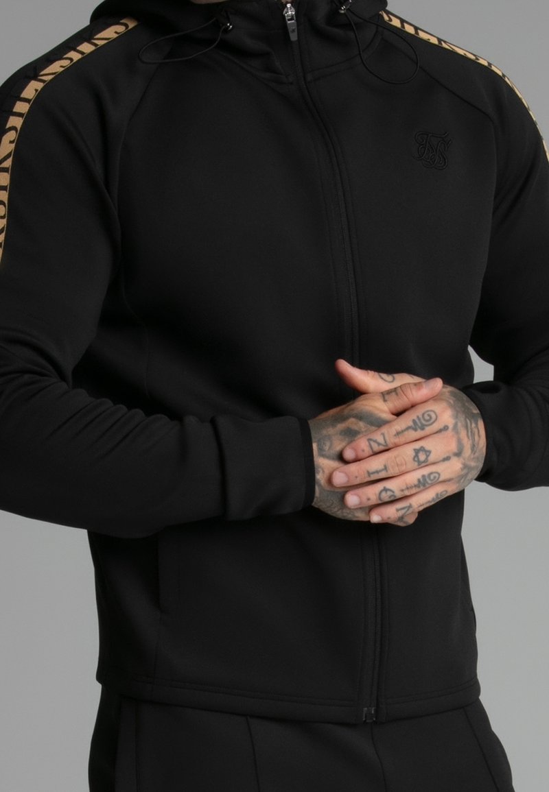 Homme portant un sweat à capuche zippé noir avec des bandes dorées sur les épaules, les mains jointes montrant des tatouages détaillés sur les doigts et les articulations.