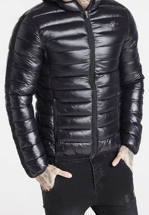 Mężczyzna w błyszczącej czarnej kurtce puchowej z zamykanymi kieszeniami i czarnych dżinsach, z rękami w kieszeniach, pokazujący tatuaże na nadgarstku i palcach.