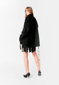 Camisa negra de gran tamaño con diseños bordados, con mangas largas y detalles de flecos, combinada con una falda negra plisada y tacones negros.