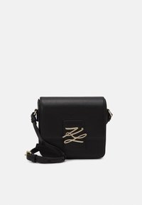 Czarna skórzana torba na ramię z klapą, ozdobiona złotym logo i regulowanym paskiem. Gładka faktura, prostokątny kształt.