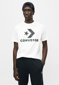 Λευκό βαμβακερό t-shirt με μαύρο λογότυπο "CONVERSE", αστέρι και βέλος· συνδυασμένο με μαύρο παντελόνι. Το μοντέλο φοράει μπλε ριγέ καπέλο.