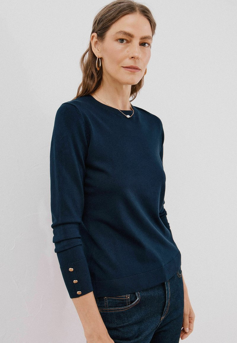 Granatowy sweter z miękkiego materiału, z długimi rękawami, z trzema dekoracyjnymi guzikami przy każdym mankiecie oraz okrągłym dekoltem.