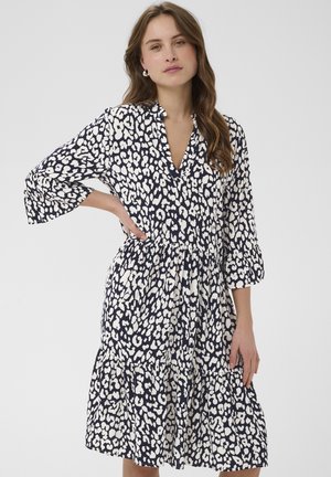 Femeie cu păr lung și ondulat, purtând o rochie alb-negru cu pete, până la genunchi, cu mâneci trei sferturi și talie încrețită, stând în fața unui fundal simplu.