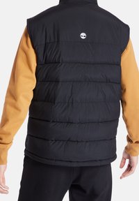 Czarna ocieplana kamizelka z widocznym poziomym przeszyciem, logo na plecach, noszona na żółtej koszulce z długim rękawem, zestawiona z czarnymi spodniami.