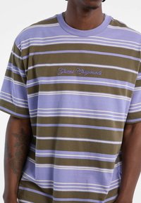 Gestreiftes T-Shirt in Olivgrün, Lavendel und Weiß. Mit lila "Guess Originals" Stickerei. Kurzärmlig, mit entspanntem Schnitt, aus Baumwollmaterial.