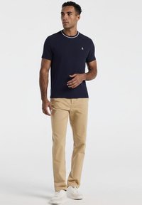 Homme portant une chemise à manches courtes bleu marine avec une bordure blanche, un pantalon beige et des baskets blanches, debout devant un fond clair uni.