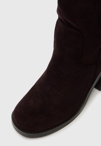 Burgundi velúr bokacipő lekerekített orral, alacsony blokk sarokkal és diszkrét varrással az oldalán. Sima textúra minimális fém kiegészítéssel.