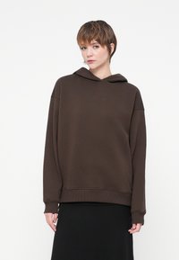 Hnedý oversize mikina s jemnou textúrou, s kapucňou na šnúrku a rebrovanými manžetami, kombinovaná s čiernou sukňou.