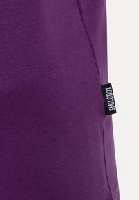 Primer plano de una tela de algodón morada con una textura suave; presenta una etiqueta de marca negra etiquetada como "SMILODON" en la costura lateral.