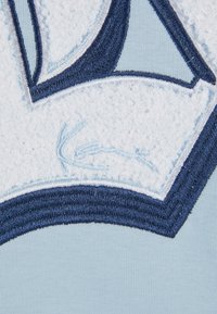 Tessuto blu chiaro con un design ricamato testurizzato bianco e blu navy. Presenta un contorno circolare e un'abbondante cucitura per i dettagli.