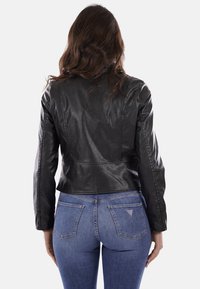 Giacca nera in pelle corta con maniche lunghe, texture liscia e dettagli di cucitura sul retro, abbinata a jeans blu con due tasche posteriori.